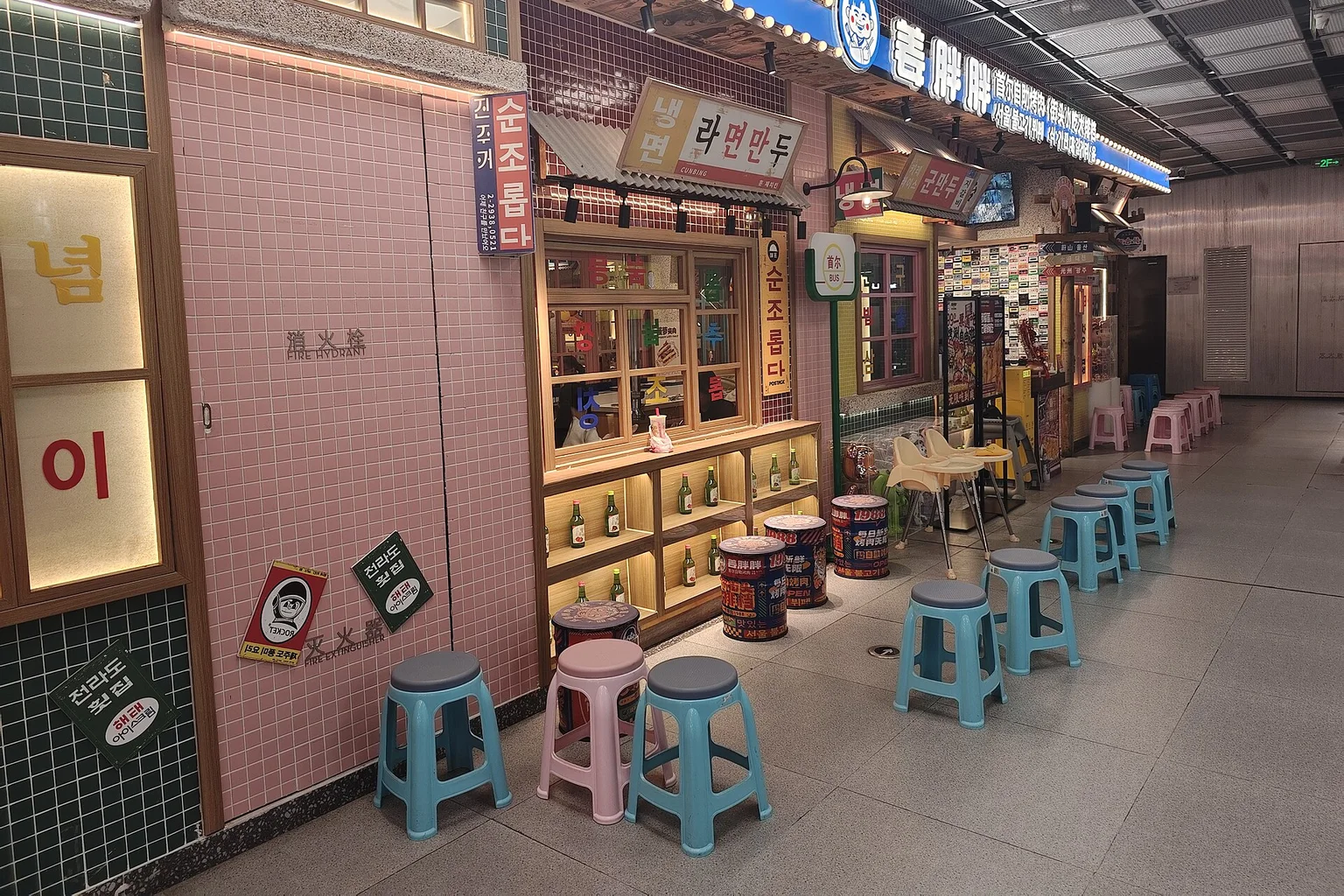 A row of Korean-style restaurant fronts with blue and pink stools in an indoor mall corridor, with no people visible.