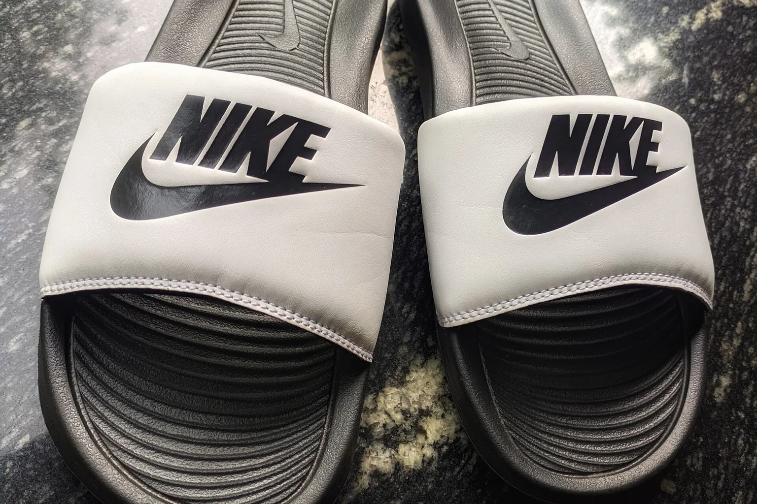 Pair of men's Nike slides with white straps and black soles on a dark speckled stone surface