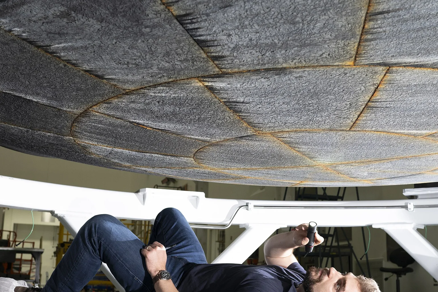 A NASA engineer lies on the floor with a flashlight, inspecting the dark underside of the Artemis II Orion heat shield.