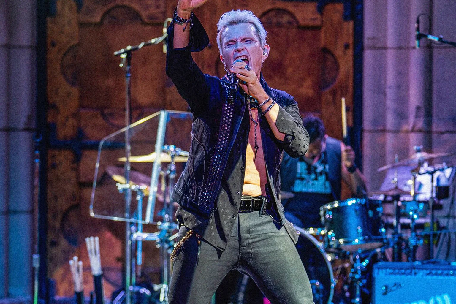 Billy Idol with silver-blond hair singing into a microphone, fist raised, with a drummer behind him on a lit stage
