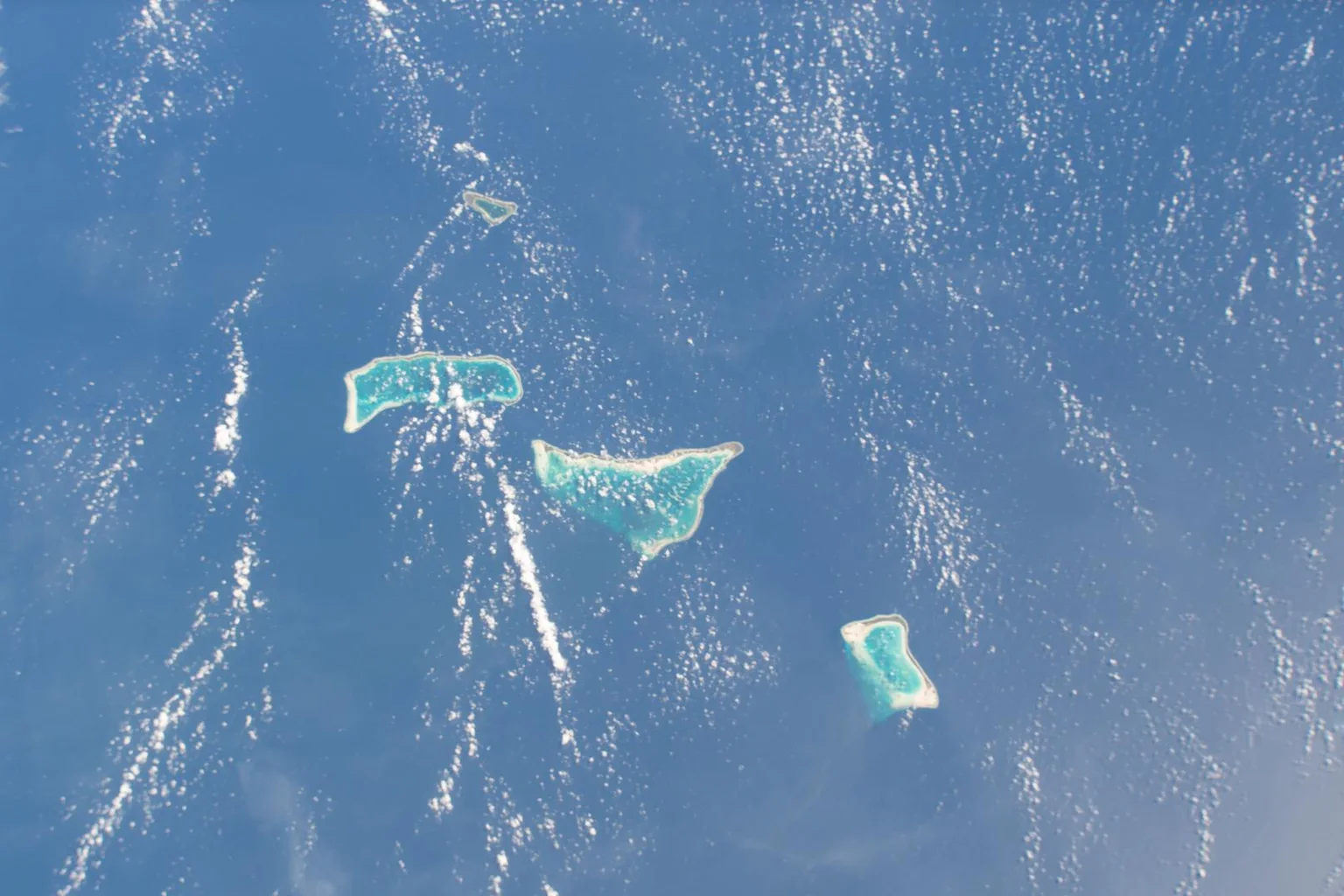 Aerial view from space of several small turquoise-lagoon islands in the Pacific Ocean, with white wave patterns on blue water.
