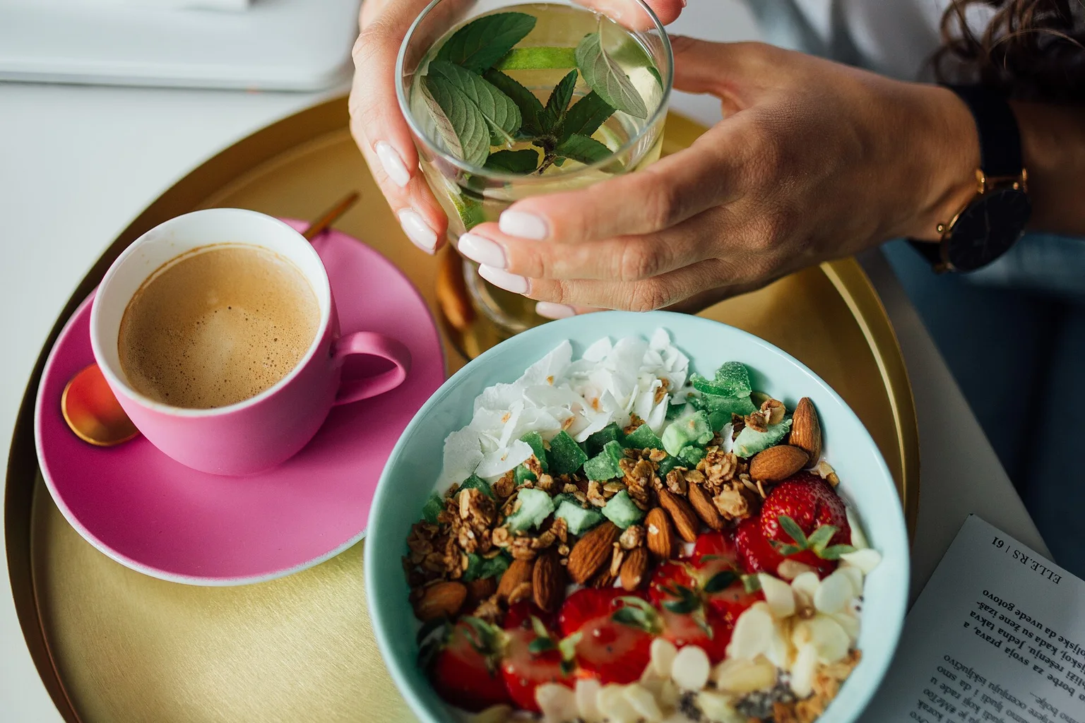 Hands holding a glass of green smoothie next to a bowl of granola topped with fruit and nuts, with a cup of coffee on a tray.