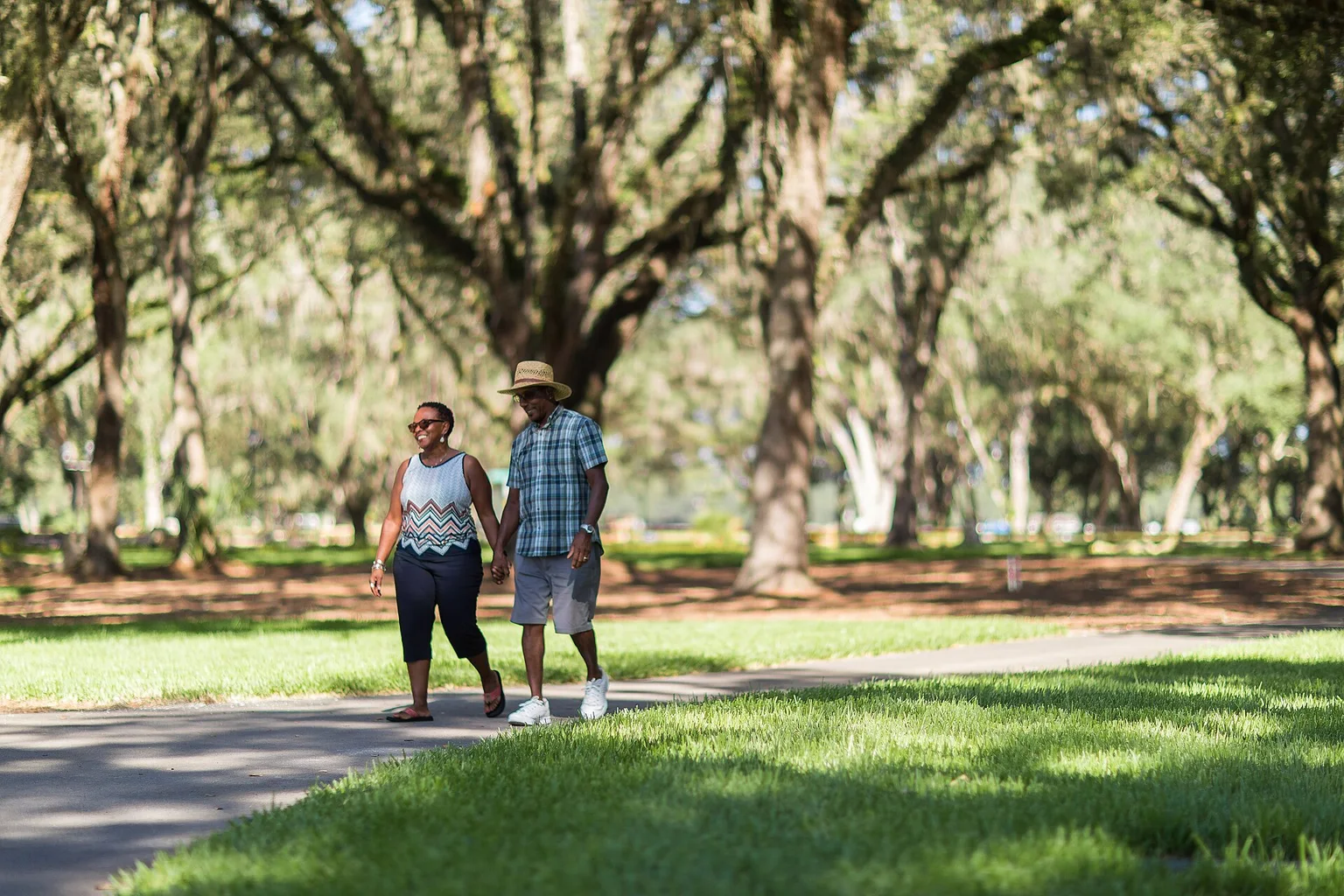 A smiling man and woman holding hands as they walk along a paved park path shaded by large oak trees.