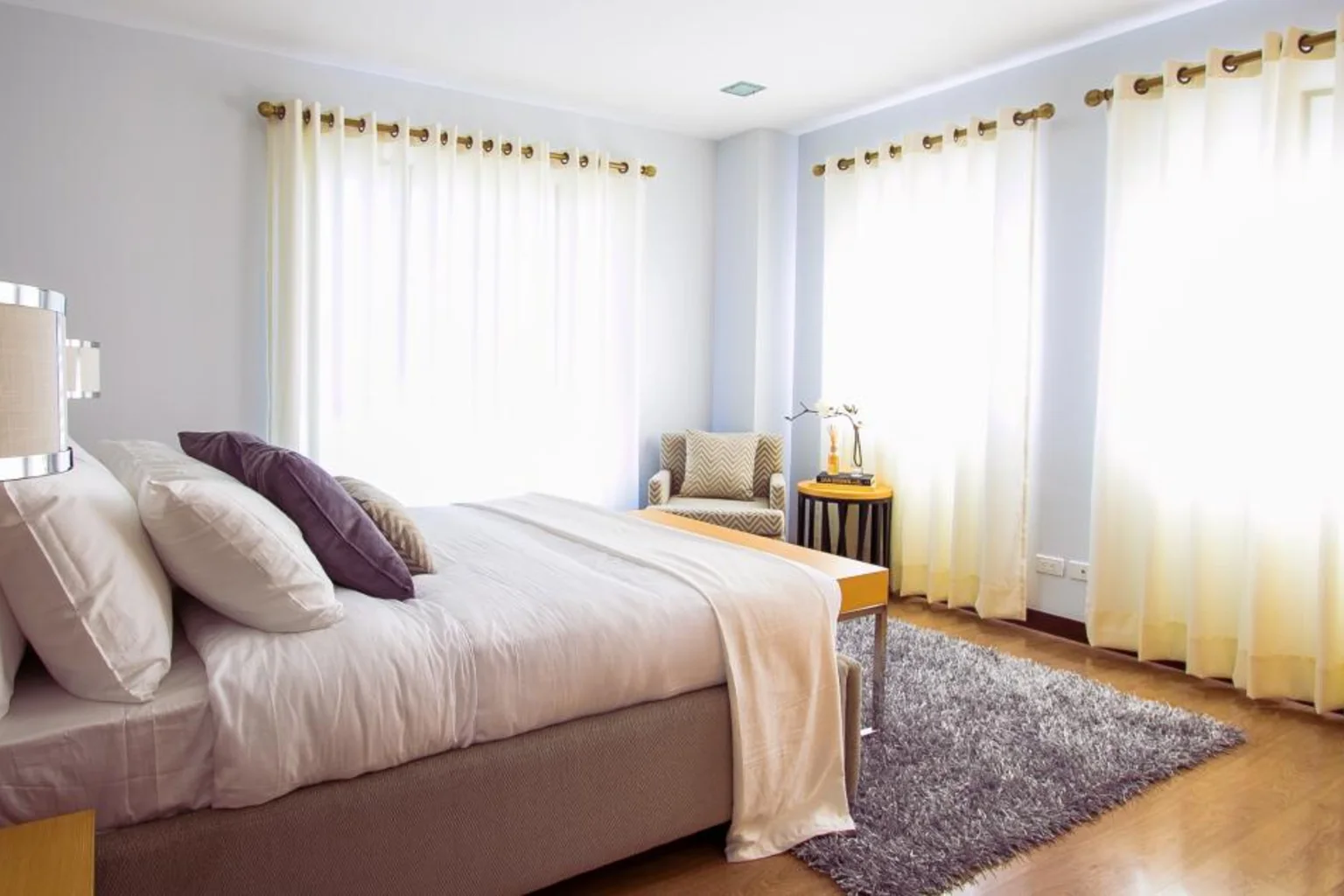 A sunlit bedroom with a bed with white and purple pillows, sheer curtains, a gray rug and a small armchair