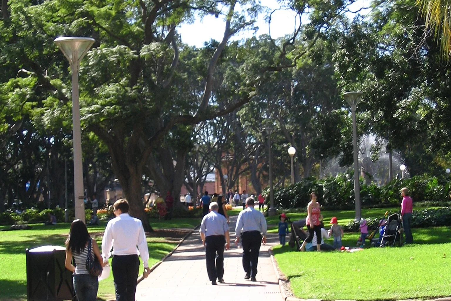 Pedestrians walk along a paved path under large green trees in a sunny city park, with families sitting on the grass nearby.