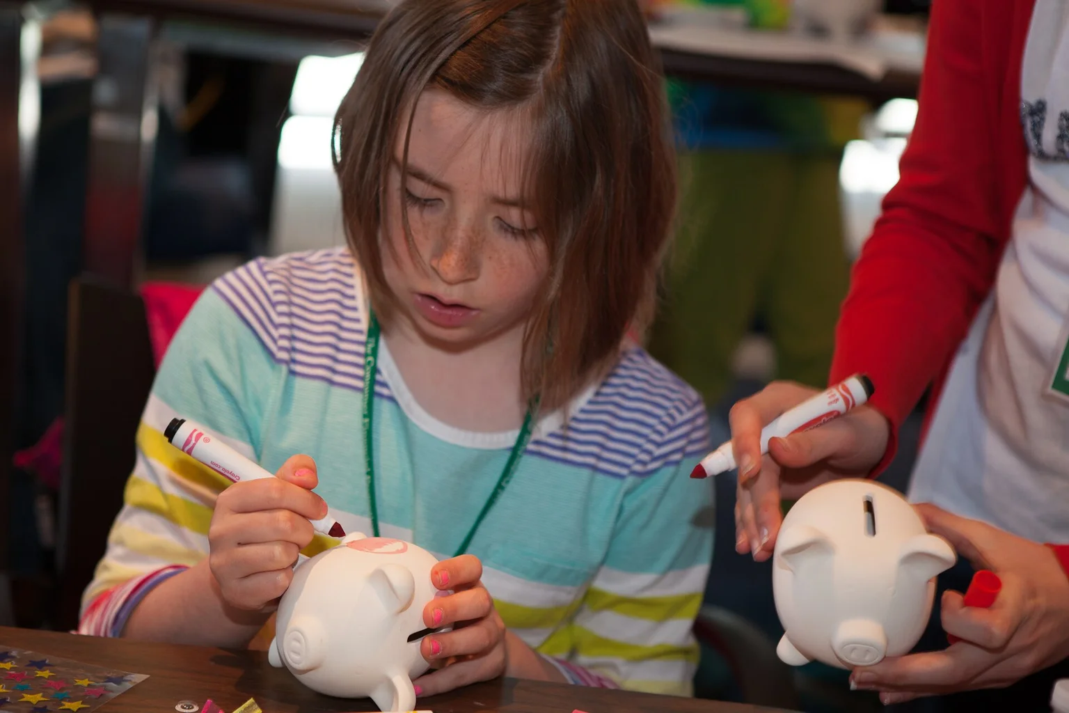 A girl with a lanyard paints a white ceramic piggy bank with a marker while an adult holds another piggy bank.