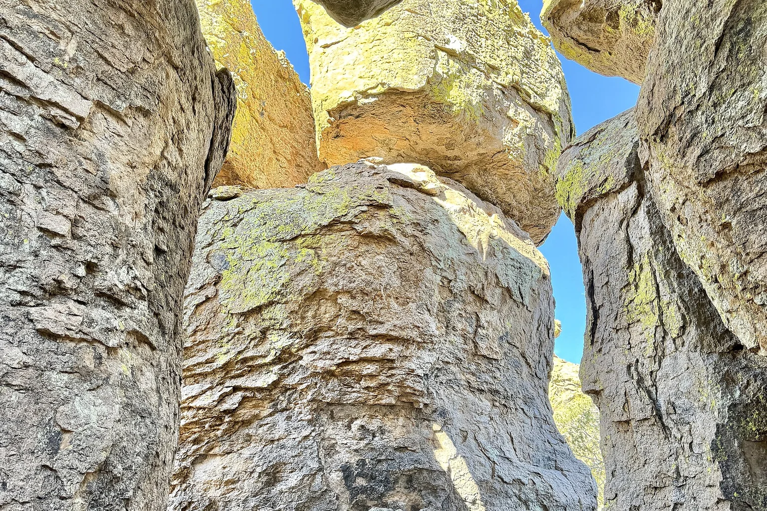 Looking up between towering volcanic rock pillars and balanced boulders, covered in yellow-green lichen, against a bright blue sky in Chiricahua National Monument.