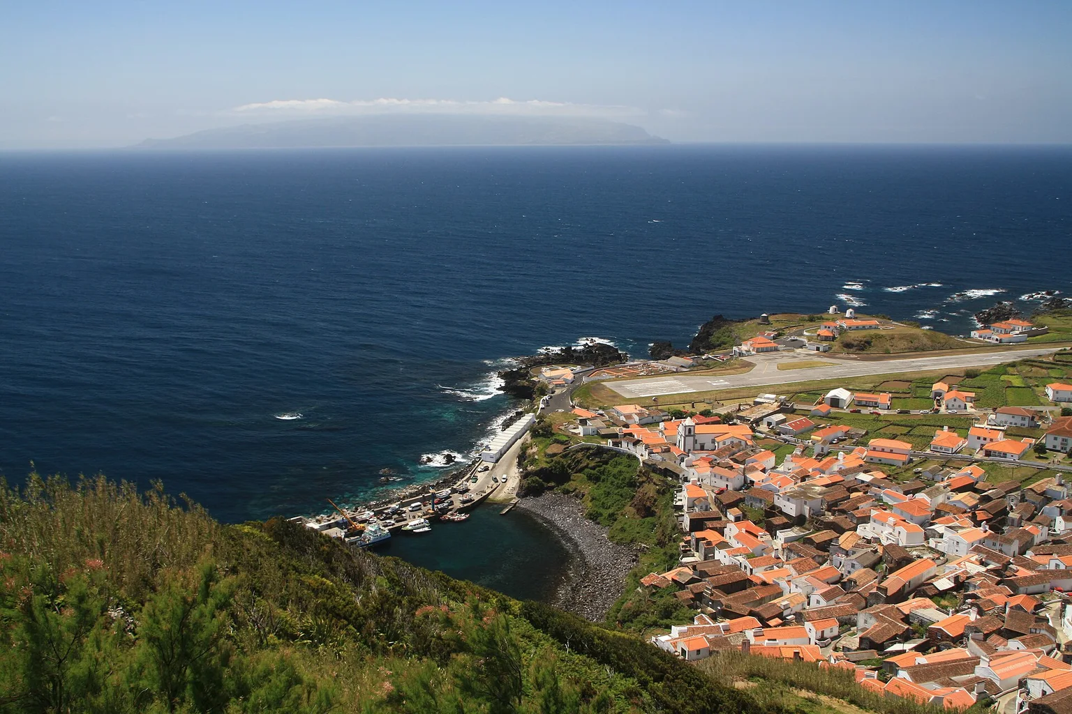 Hillside view over the orange-roofed houses of Vila do Corvo on Corvo island, with the small harbour, airstrip and deep blue Atlantic beyond.