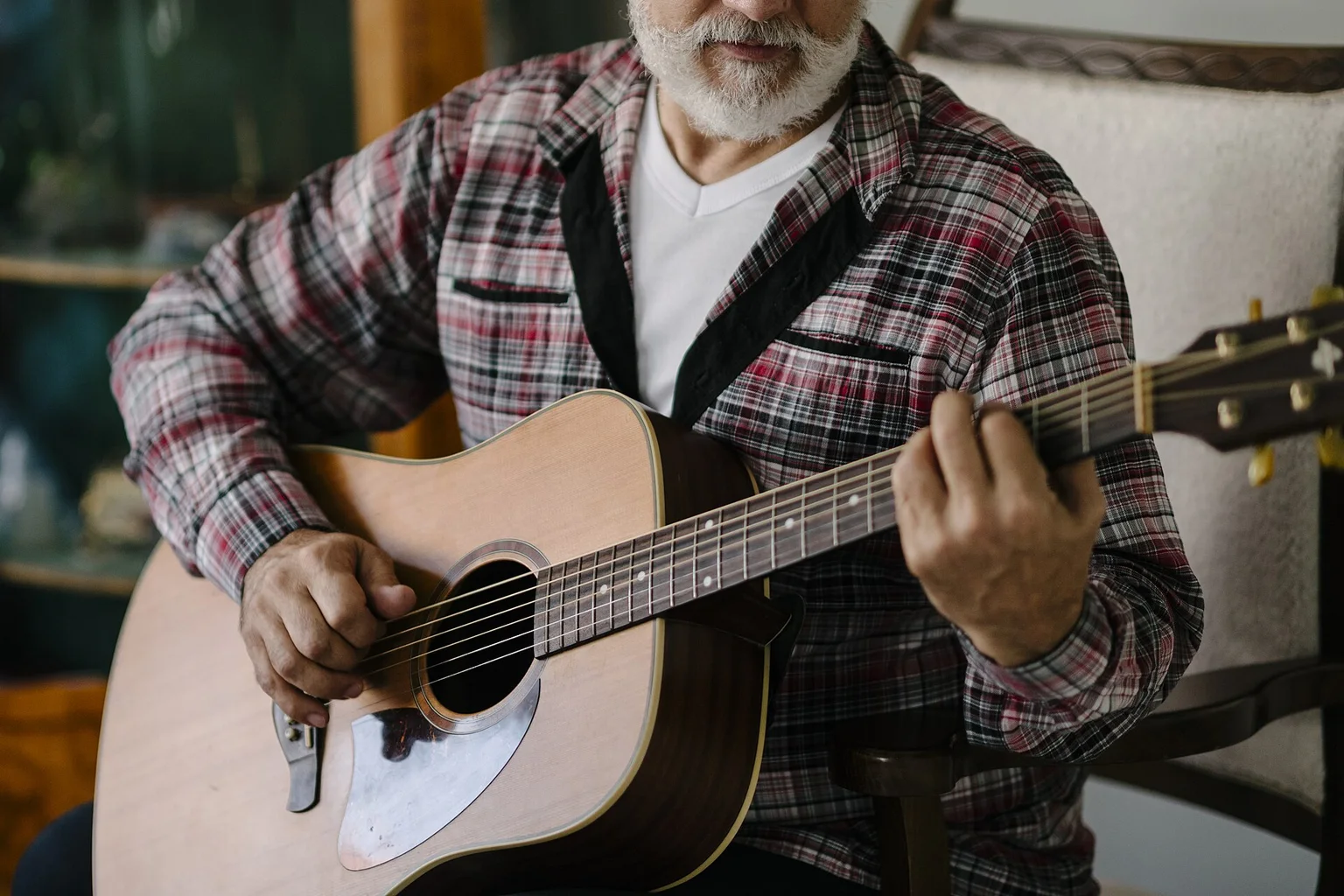 Close view of an older bearded man in a plaid shirt strumming an acoustic guitar while sitting in a chair