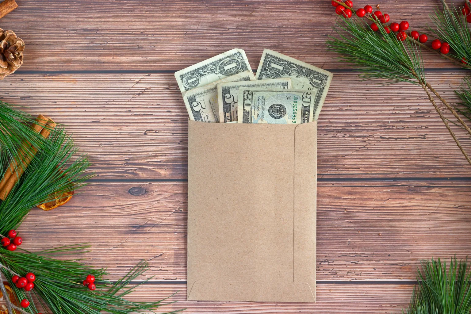 A brown paper envelope with several US dollar bills tucked into its top, on a rustic wooden surface decorated with pine sprigs and red berries.