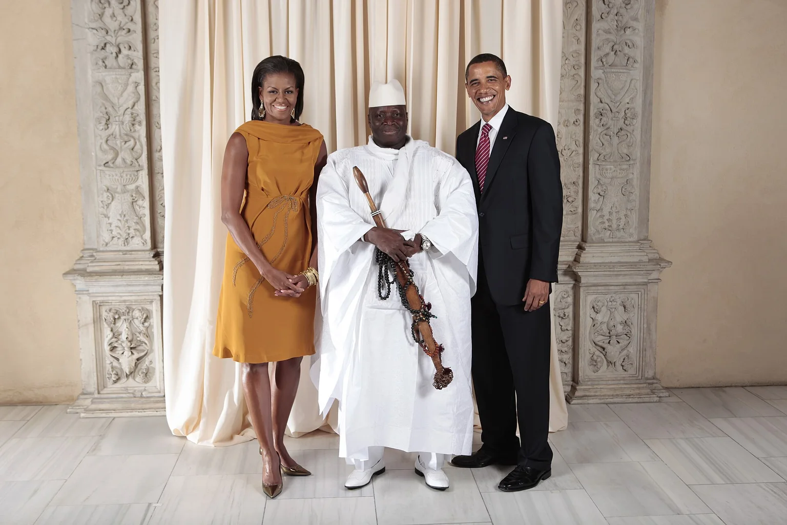 Yahya Jammeh in white robes and cap holding a ceremonial staff, standing between Michelle Obama and Barack Obama in front of a cream curtain.