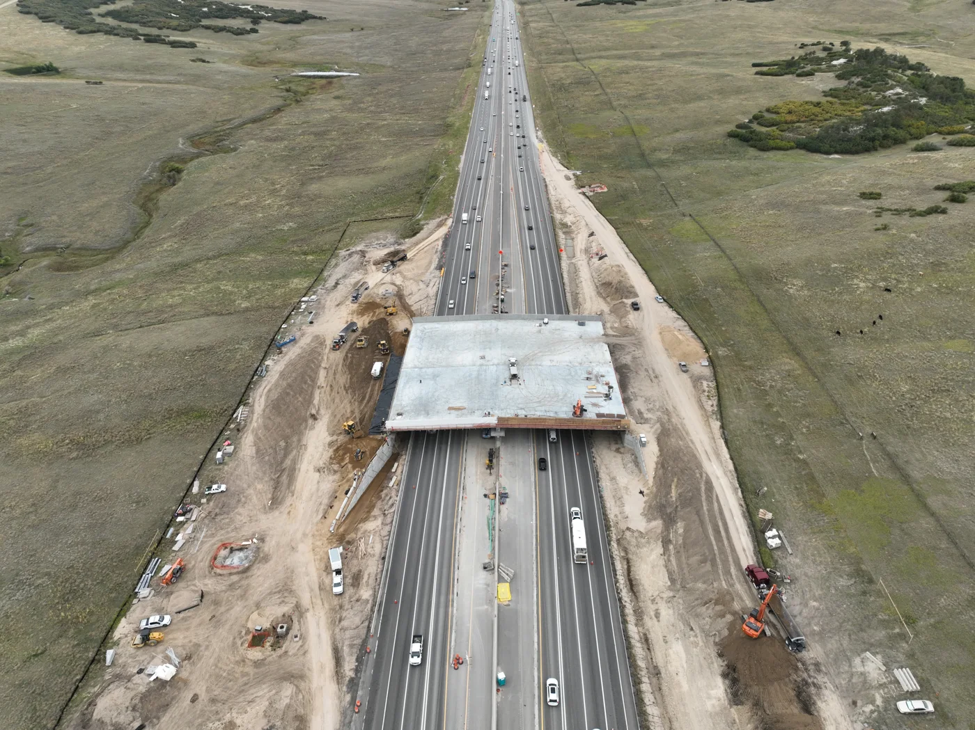 Aerial view looking north along Interstate 25, with the bare concrete deck of the wildlife overpass spanning all lanes and construction equipment along the shoulders.