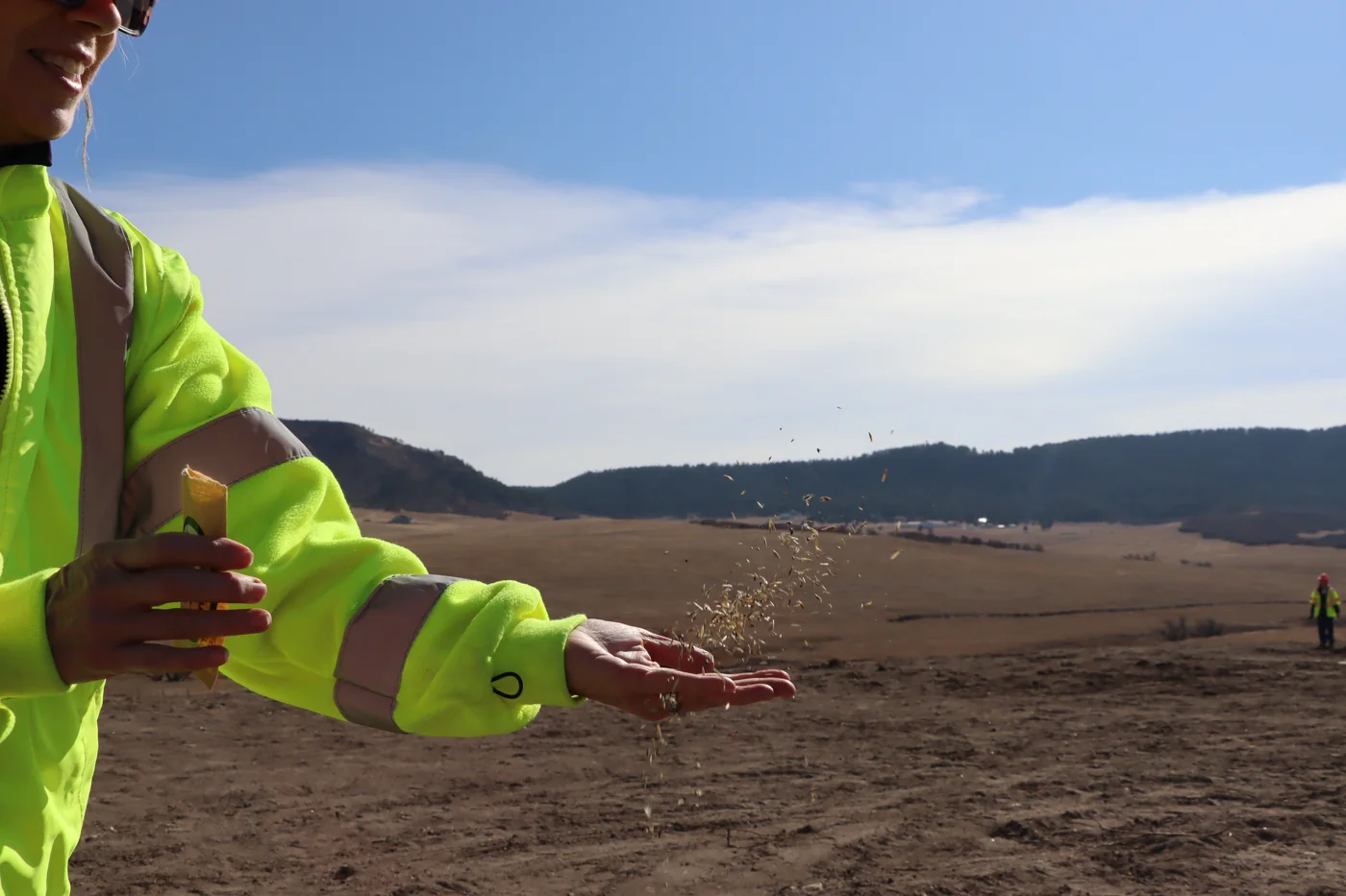 A CDOT worker in a bright yellow safety jacket scatters native seed by hand across the bare soil of the overpass.