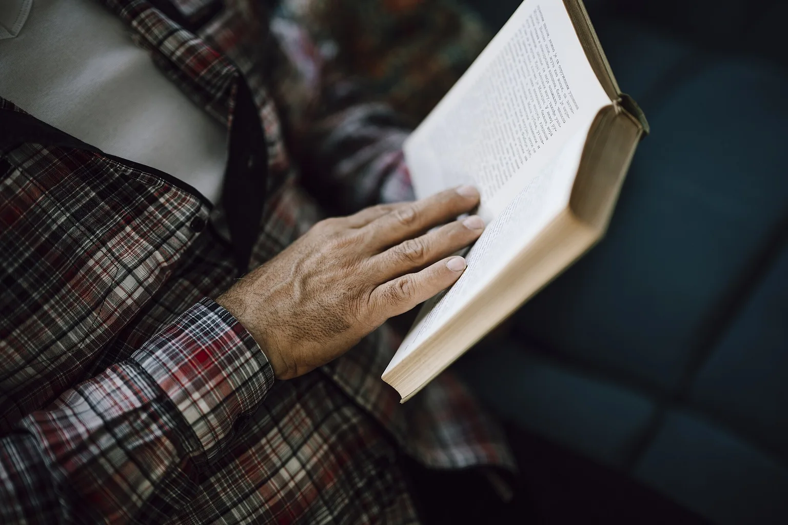 Close-up of a person in a plaid shirt reading an open book, one hand resting on the page