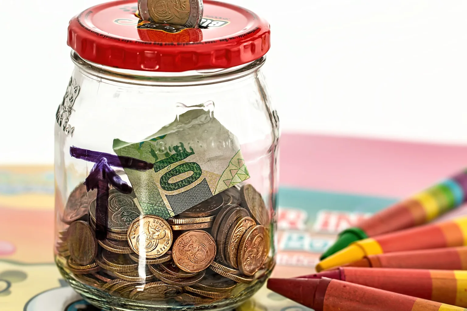 Glass jar with a red lid and a coin being dropped in, holding coins and a folded banknote, with crayons beside it