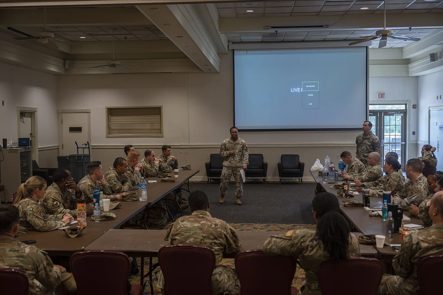 A uniformed facilitator stands at the front of a conference room while service members seated at tables listen to a discussion session.