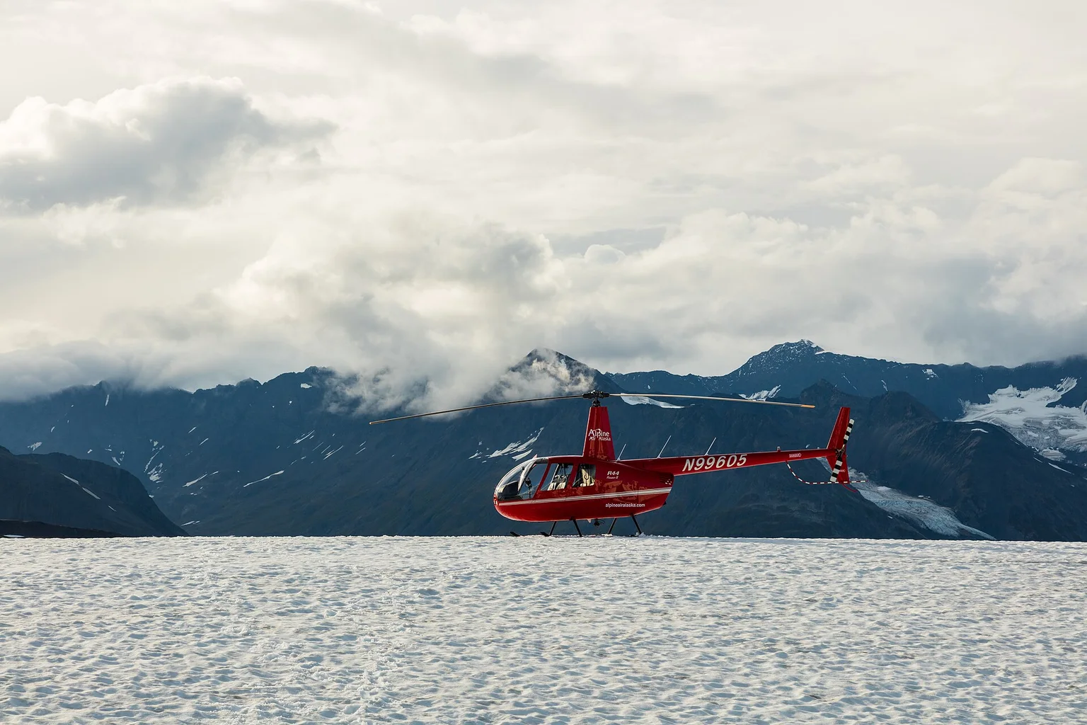 Red Robinson R44 helicopter sitting on a snowfield with cloud-covered Alaskan mountains in the background