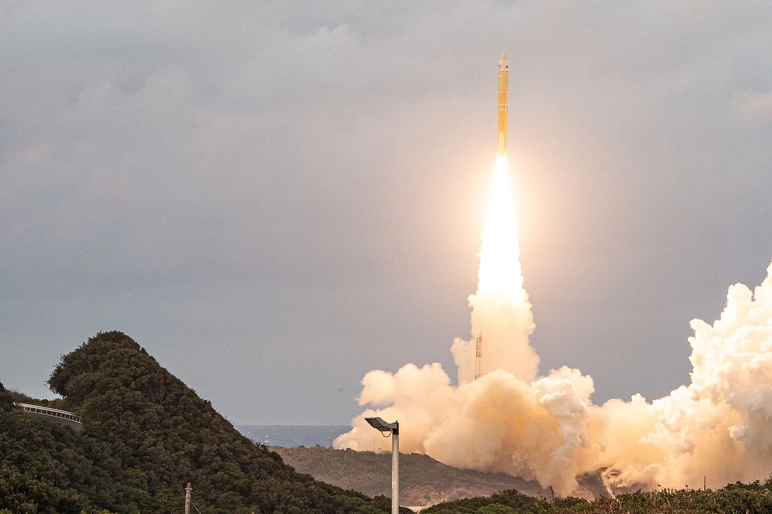 A golden-yellow H3 rocket climbs on a bright plume of fire and smoke above green hills, with a small group of spectators in the foreground.