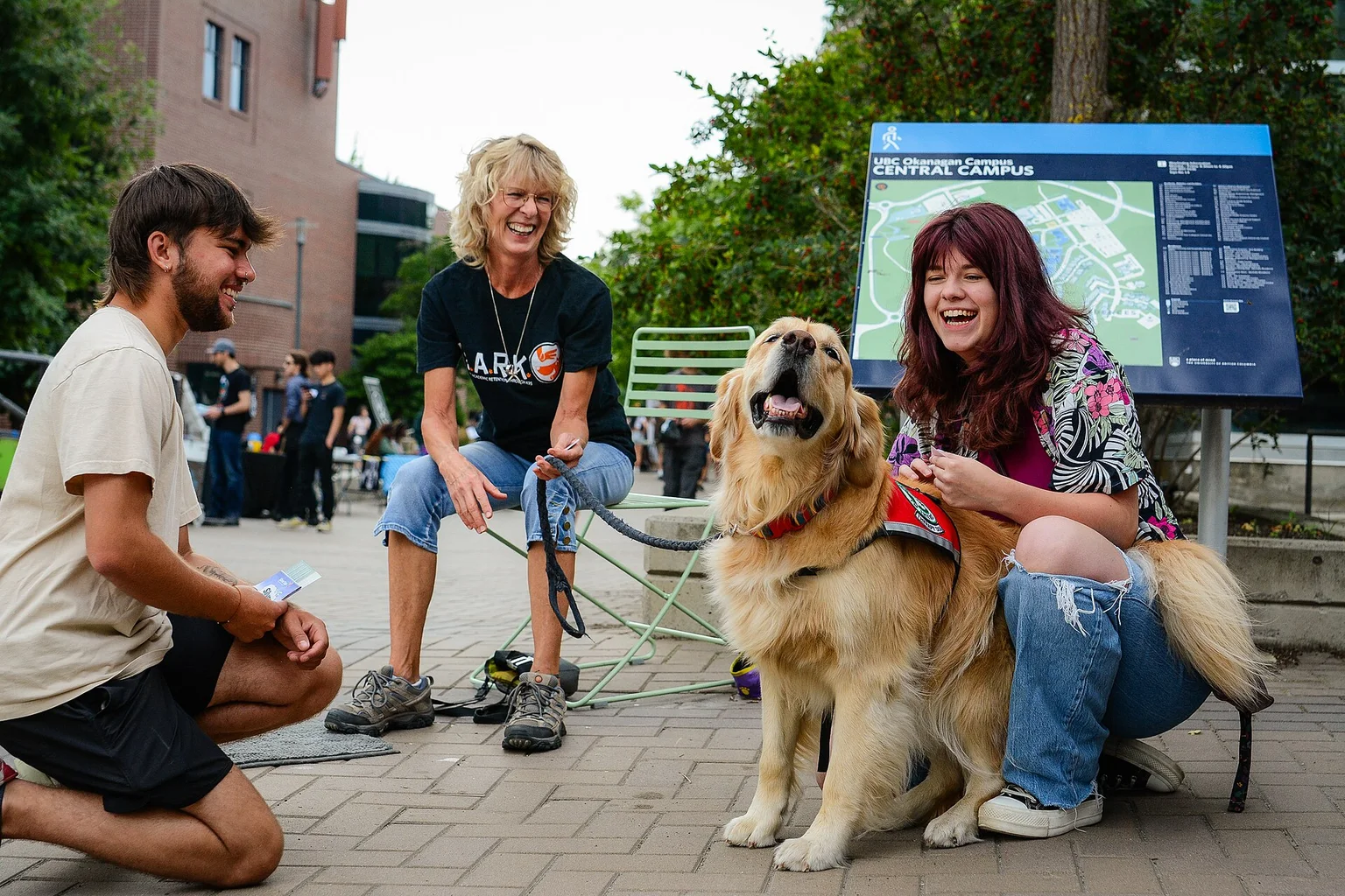 A golden retriever therapy dog in a red vest being petted by laughing students and a volunteer handler on a campus path