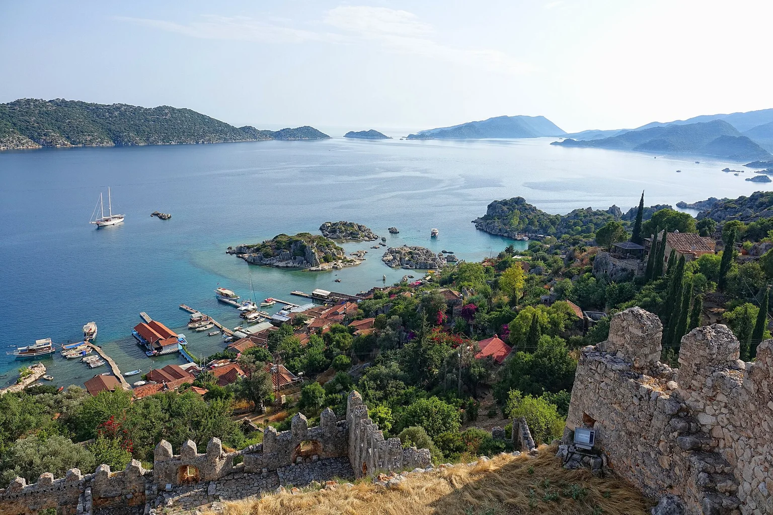 Elevated view over a turquoise bay with small islands, boats at a village harbor and old fortress walls in the foreground.