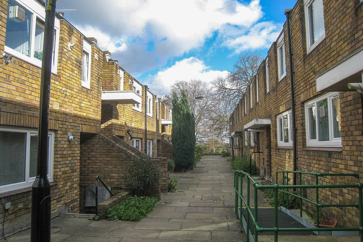 A paved pedestrian lane between two rows of yellow-brick houses on Cressingham Gardens estate under a blue sky with clouds