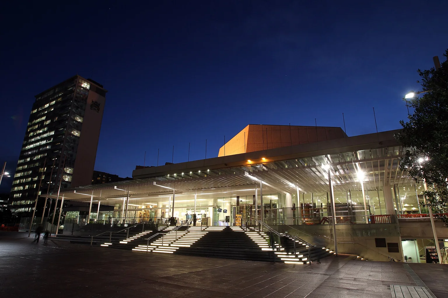 The glass-fronted Aotea Centre in Auckland under a deep blue night sky, with illuminated steps leading up to the entrance and a council tower at left.