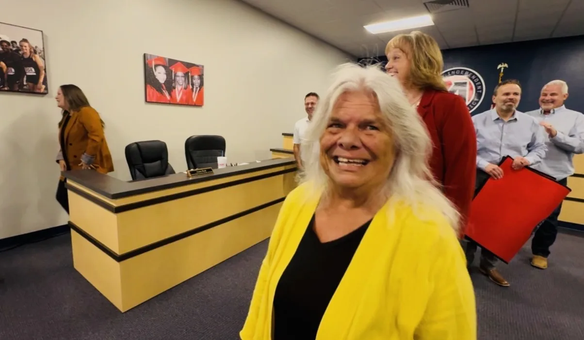 Marilyn Hampton in a yellow cardigan smiling in the Aubrey ISD board room, with a woman holding a red carpet behind her