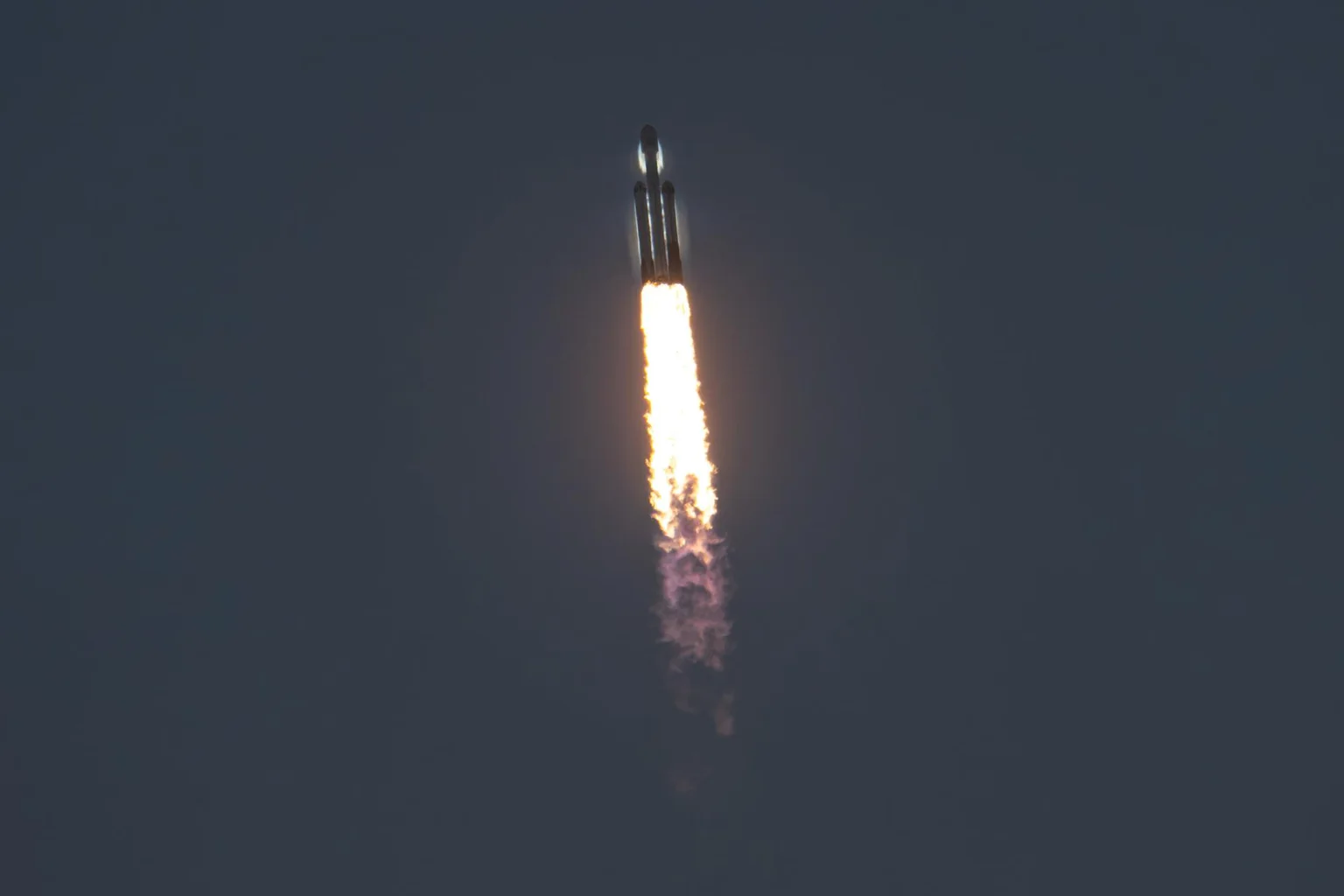 A Falcon Heavy rocket climbs through a dark sky with a bright orange exhaust plume, carrying the Roman Space Telescope.