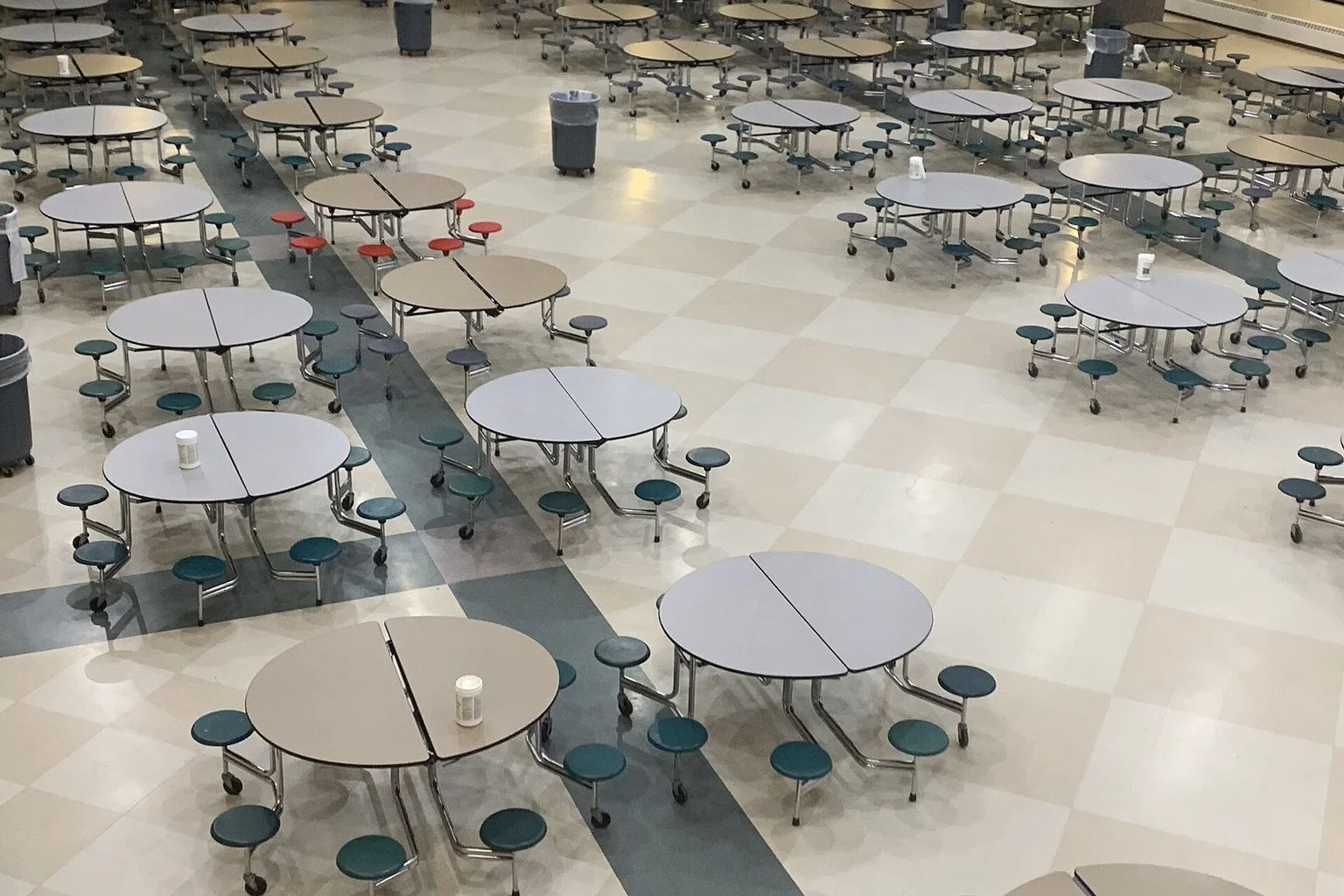 A large empty school cafeteria with round gray tables, teal stools and a checkered floor, viewed from above.