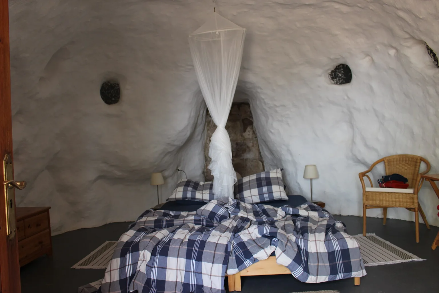 Bedroom in a white-walled cave dwelling with a double bed, plaid bedding, a mosquito-net canopy and two bedside lamps