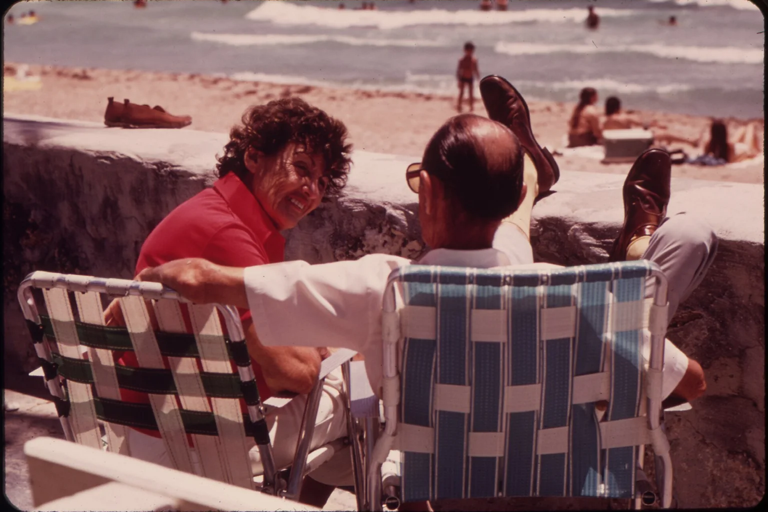 Two older people sit in folding lawn chairs on a beach wall, talking, with the ocean and beachgoers behind them.