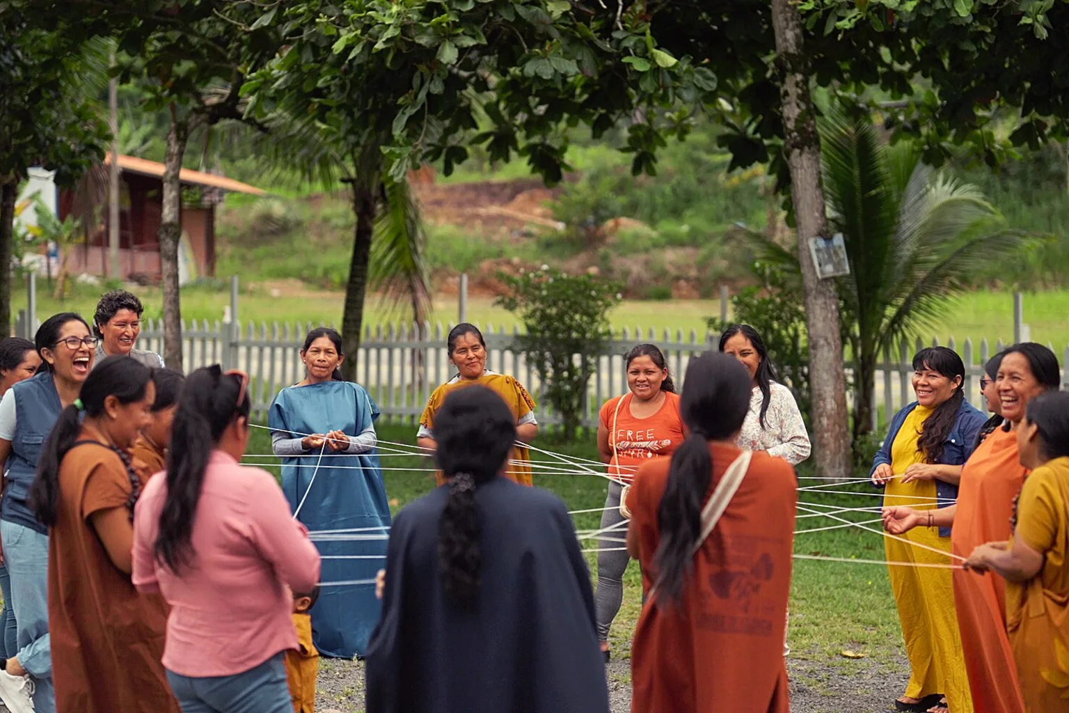 A circle of smiling Asháninka and Yanesha women in colorful traditional tunics hold strings between them during an outdoor workshop, with trees and green hills behind.