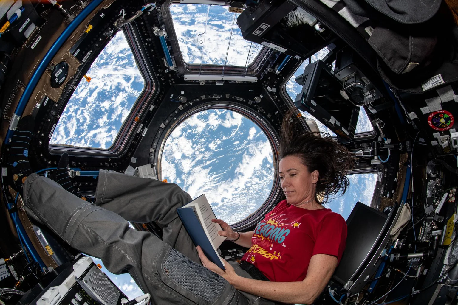 A woman floats in the space station's domed window module, reading a book with Earth and clouds visible behind her.