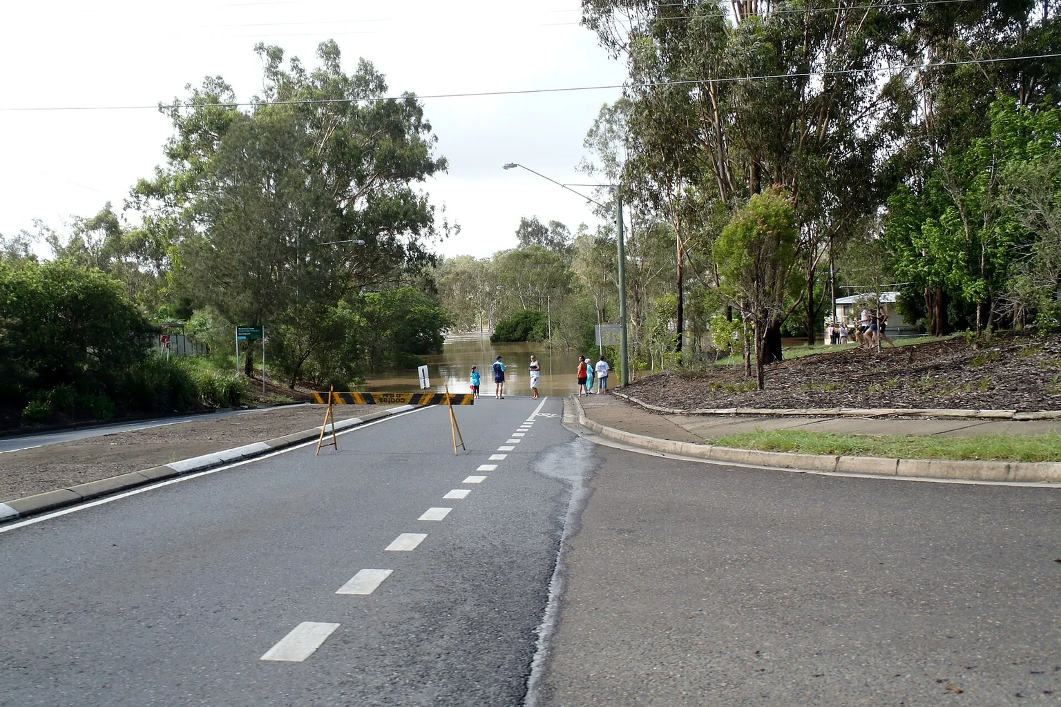 A road closed by barriers where brown floodwater covers the road, with a few people standing back and gum trees on both sides