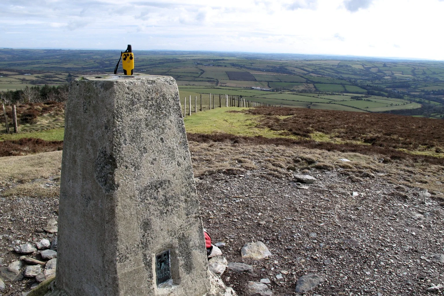 A concrete trig pillar with a yellow GPS unit on top, on the Frenni Fawr hilltop, with green fields stretching into the distance