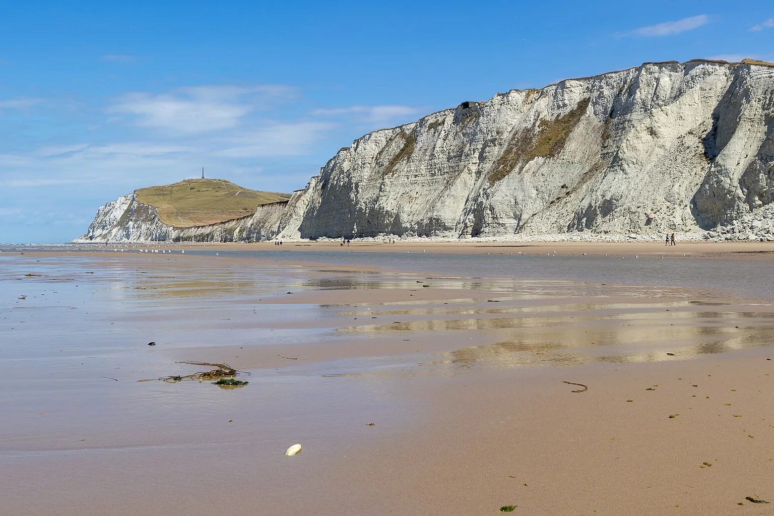 Chalk cliffs of Cap Blanc-Nez on the French coast, with a monument-topped hill beyond a wide sandy beach under blue sky.