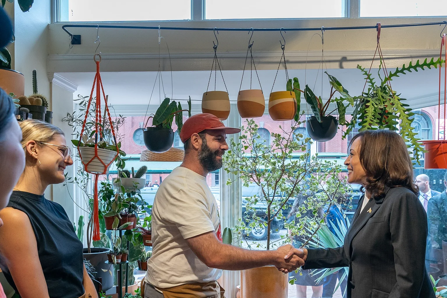 A bearded shop owner in an apron shakes hands with a woman in a dark blazer inside a plant shop filled with hanging and potted plants.