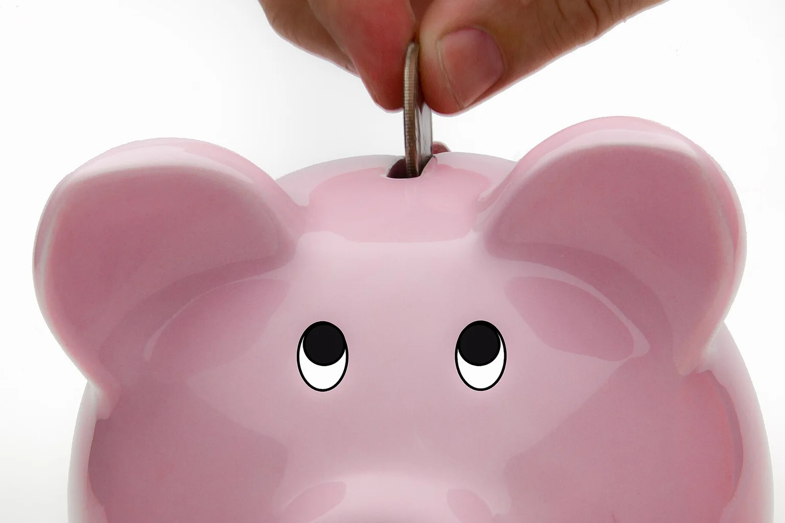 Close-up of a hand putting a coin into the slot of a pink ceramic piggy bank.