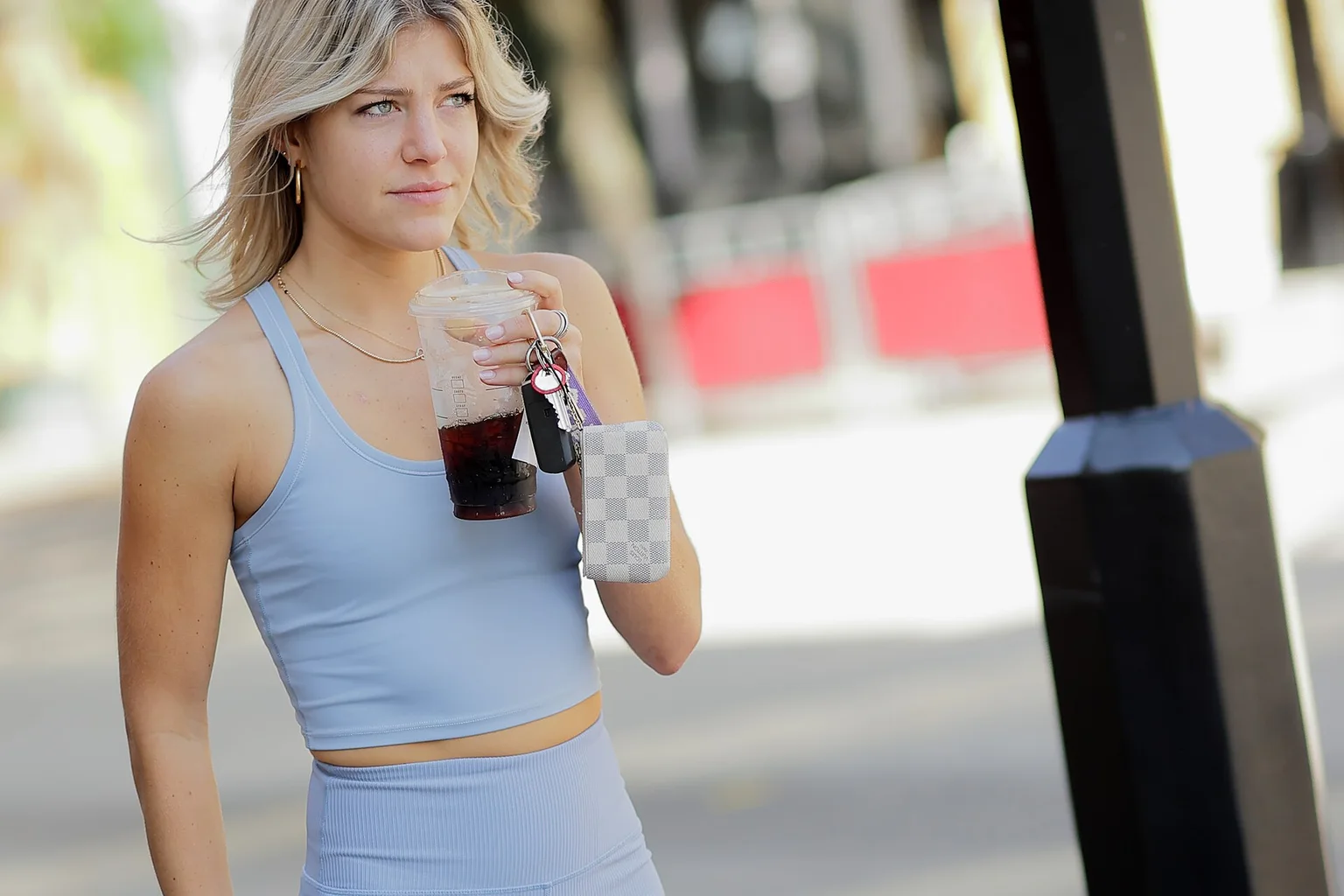 A young woman with wavy blonde hair wearing a light blue crop top and matching bike shorts, holding a drink and walking on a city sidewalk.