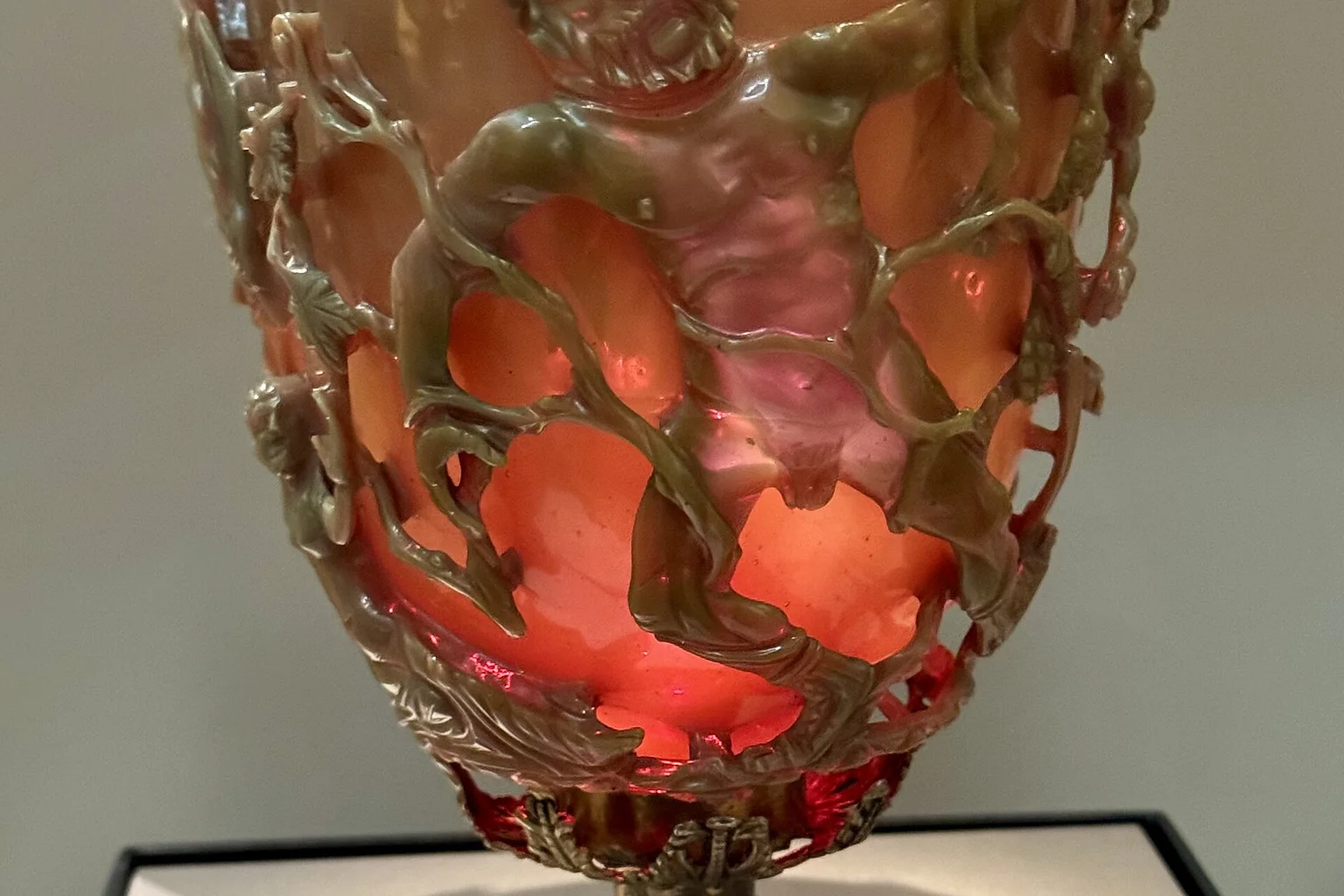 The Roman Lycurgus Cup on display at the British Museum, its carved glass figures glowing ruby red with backlighting, set in a gilded silver rim and foot.