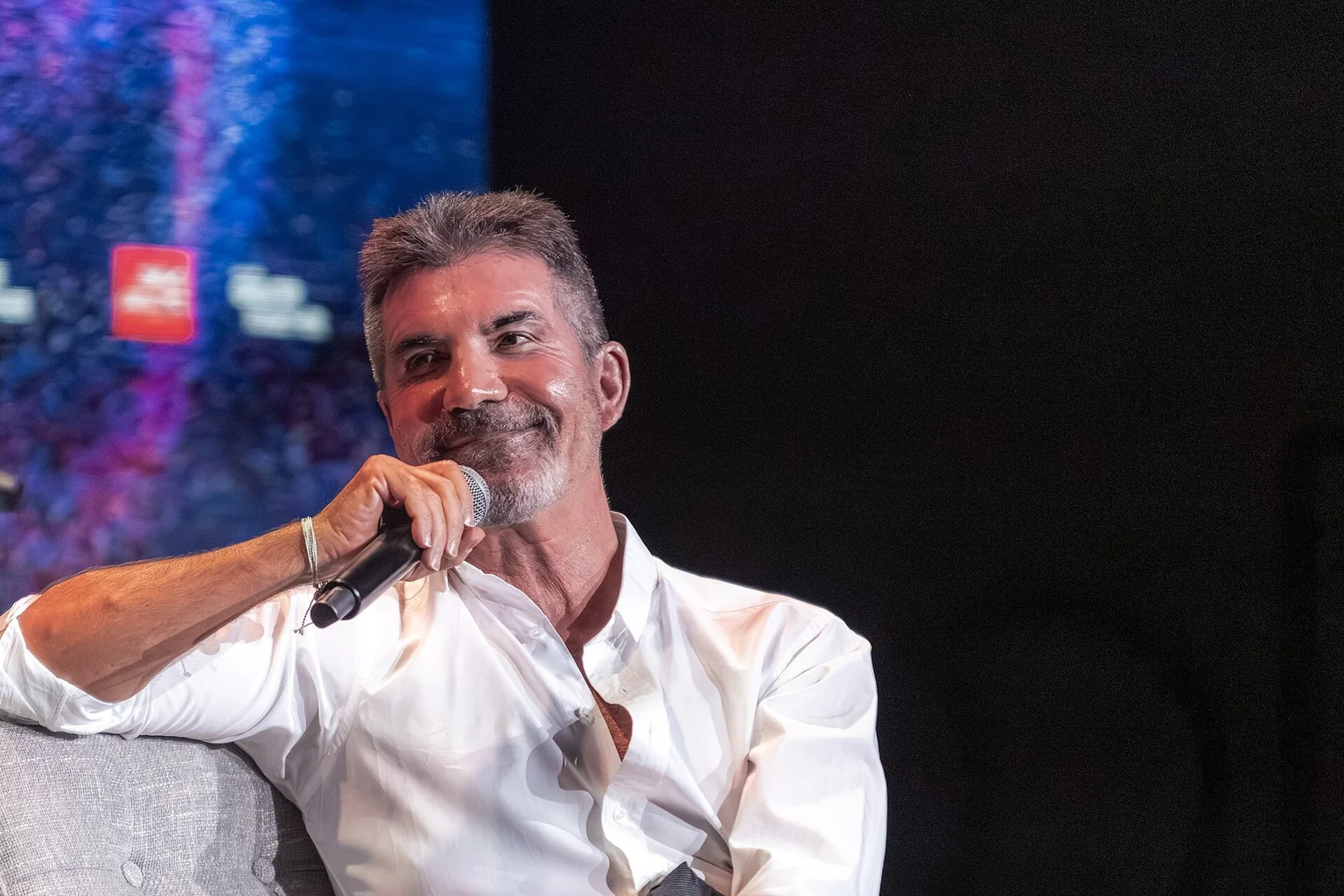 Simon Cowell, wearing a white shirt, sits in a grey armchair on stage, holding a microphone and smiling.