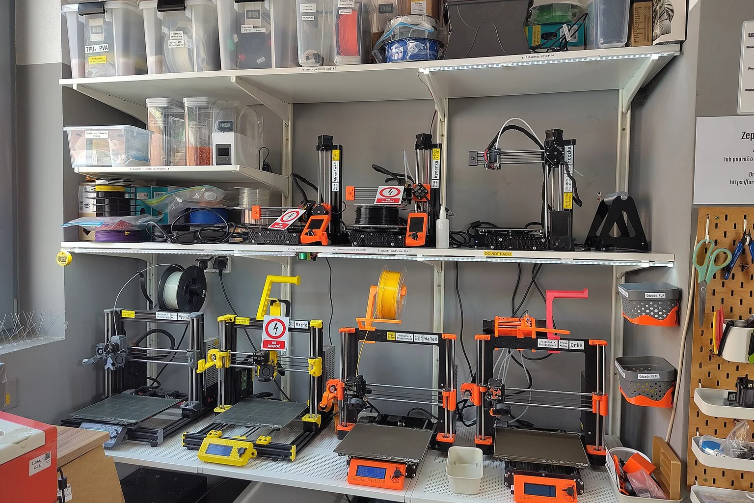 Two shelves holding several desktop 3D printers, with spools of filament and storage boxes above them.