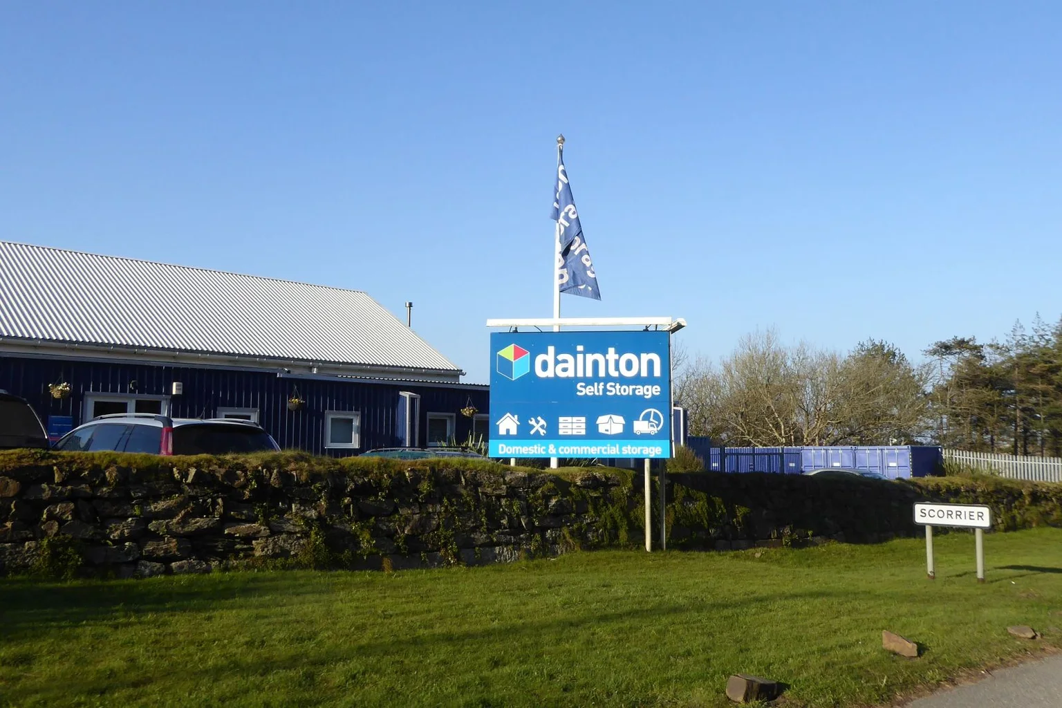 Dainton Self Storage facility with a large blue sign and blue storage containers behind a stone wall