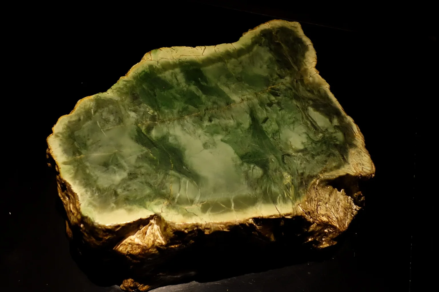A rough block of raw pounamu greenstone with a cut face showing green and pale patterns, lit against a dark background.