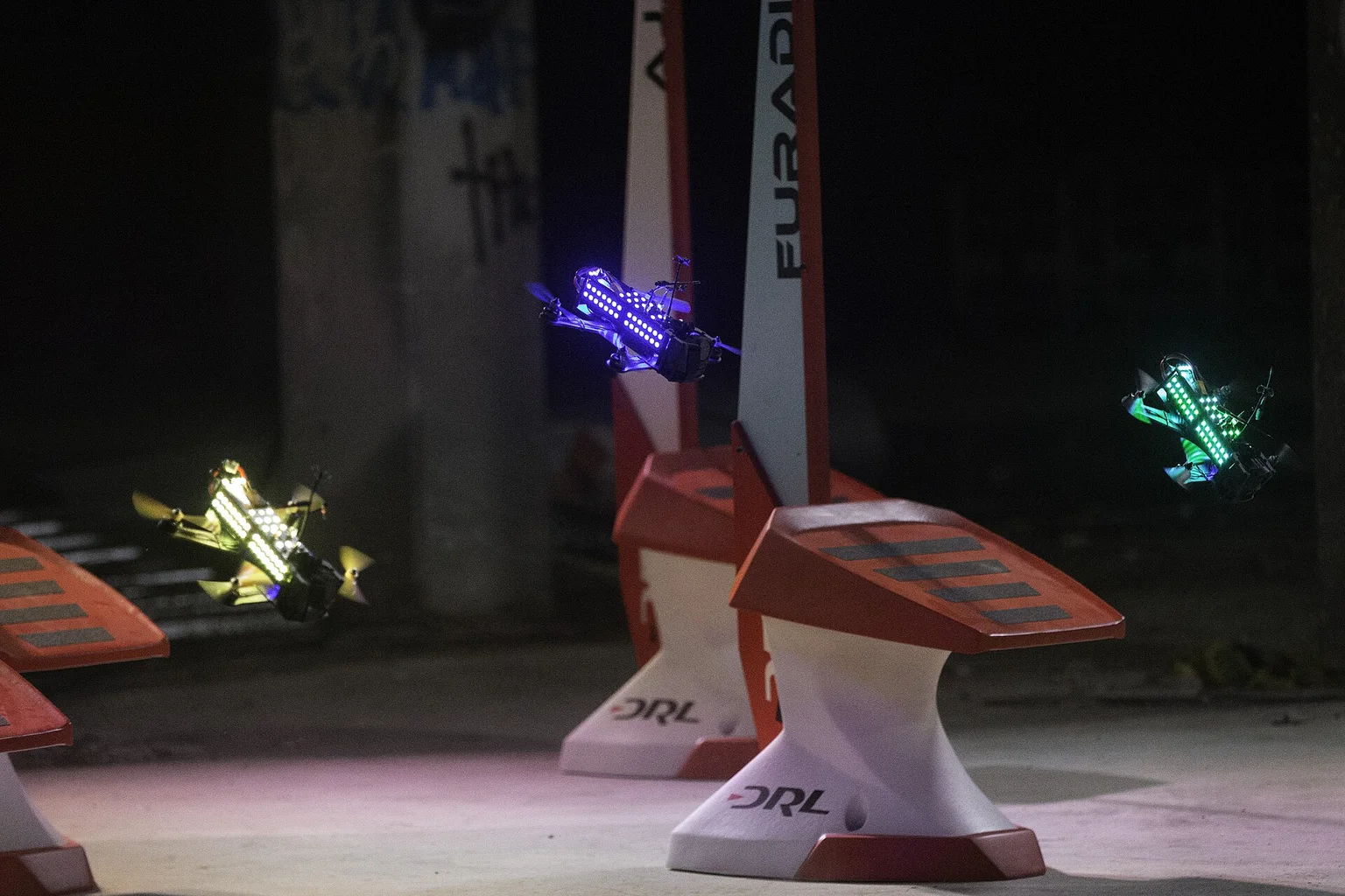 Three racing drones with glowing blue, yellow and green lights flying past race gates in a dark venue