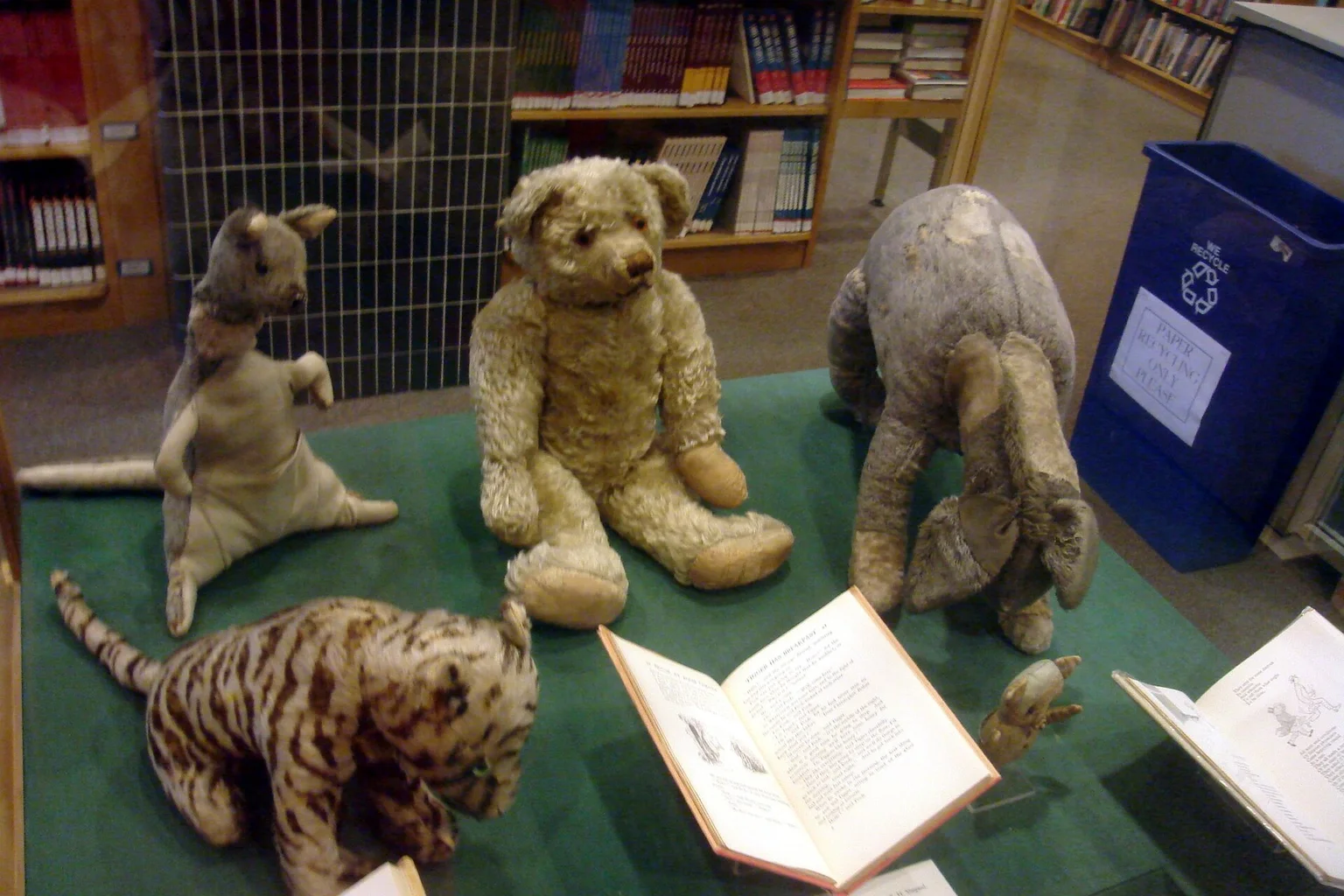 Worn vintage stuffed toys of Pooh, Eeyore, Kanga, Tigger and Piglet on a green display table beside an open book in a library.