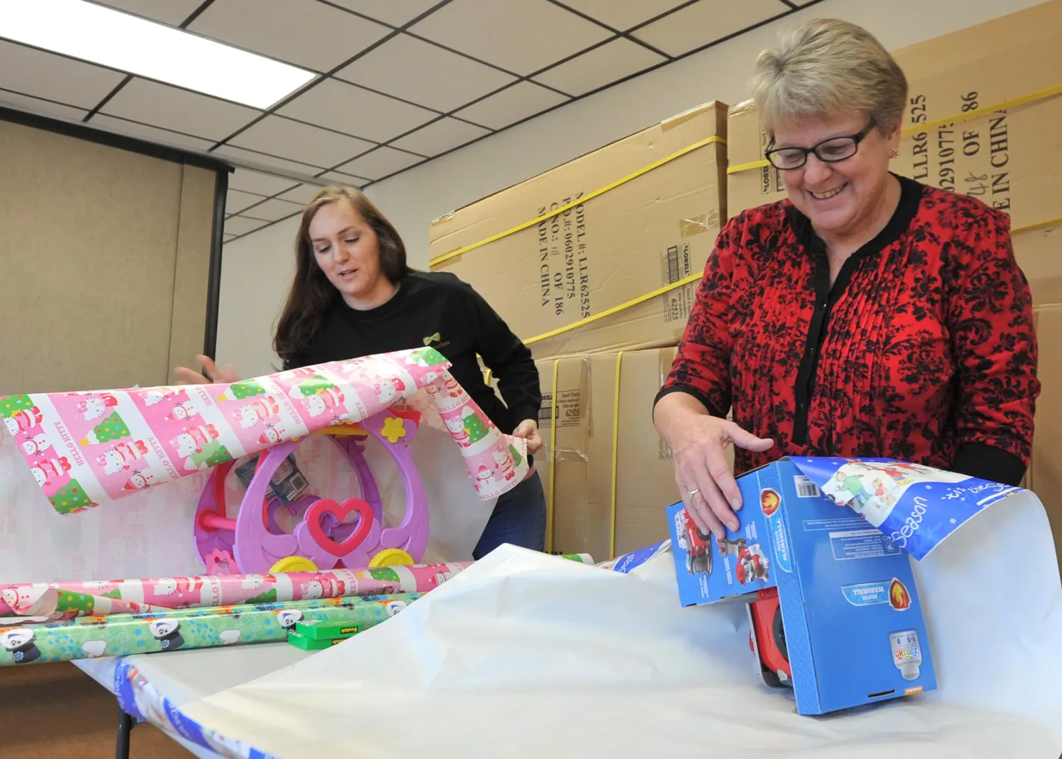 Two women wrapping toys in colorful paper at a long table.