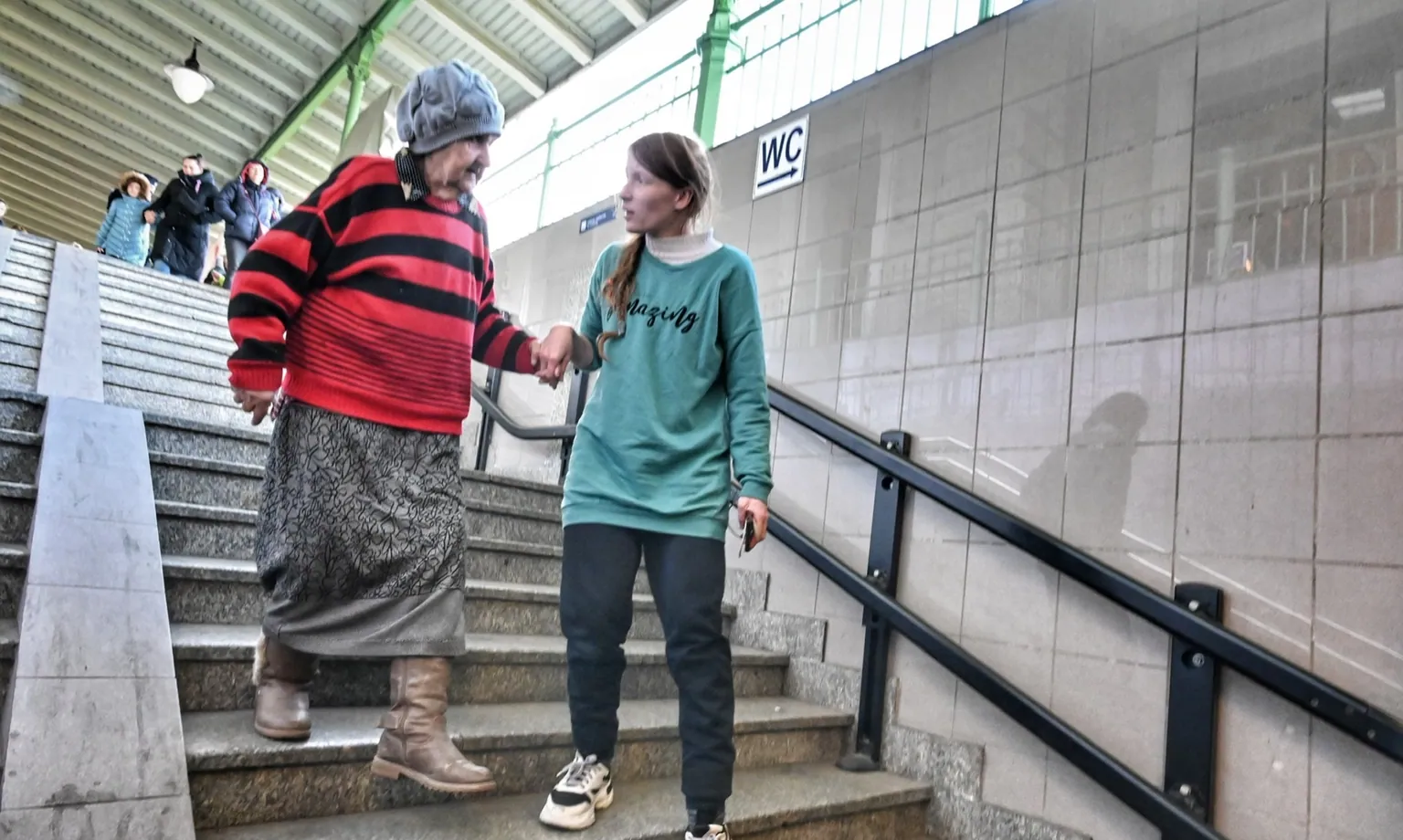 A young woman in a green sweatshirt holds the hand of an older woman as they walk down station stairs.