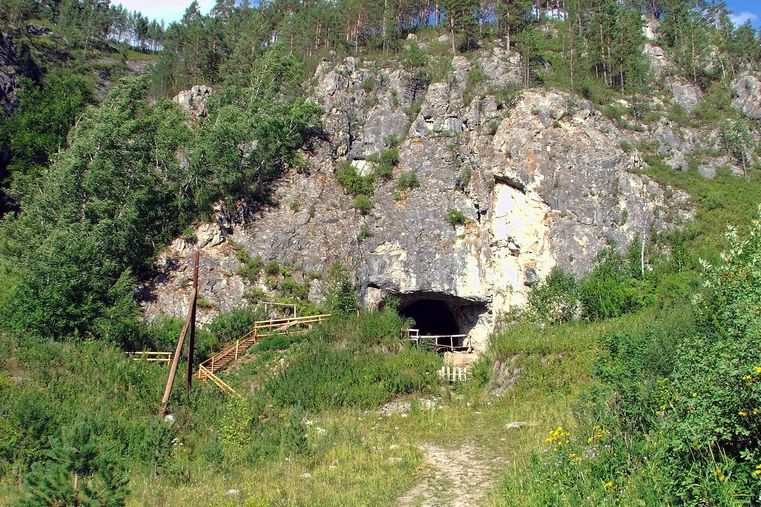 Rocky hillside with trees above the dark entrance of Denisova Cave, with a wooden staircase and a grassy path in the foreground