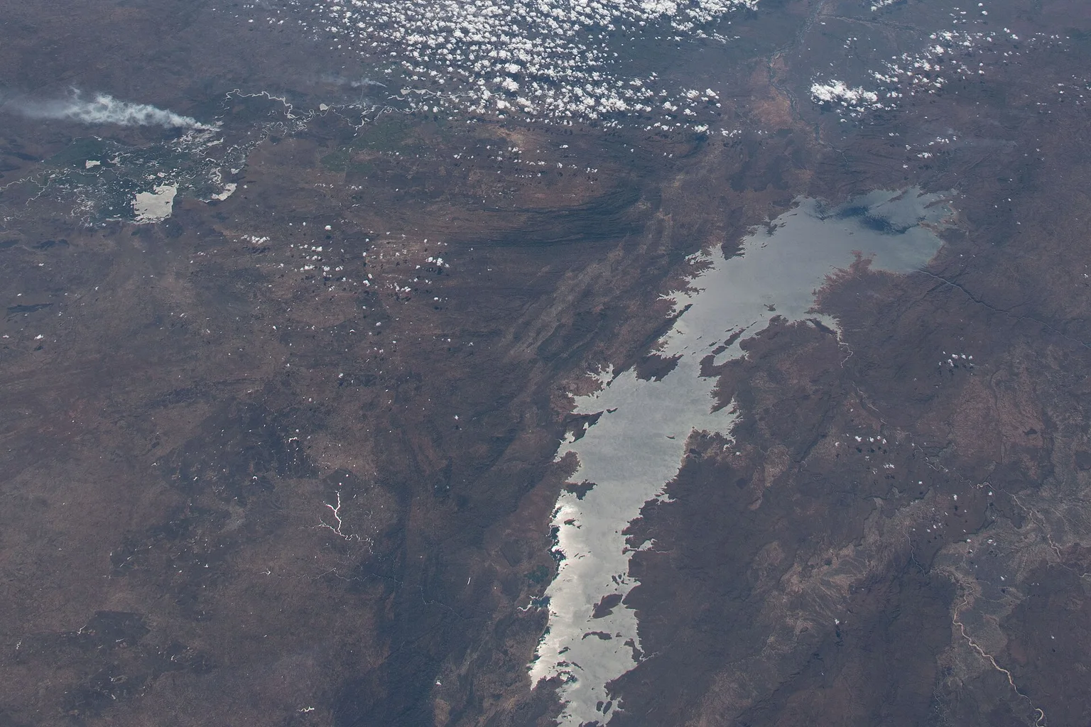 Photo taken from the International Space Station of Lake Kariba, a long narrow lake among brown land and scattered clouds.